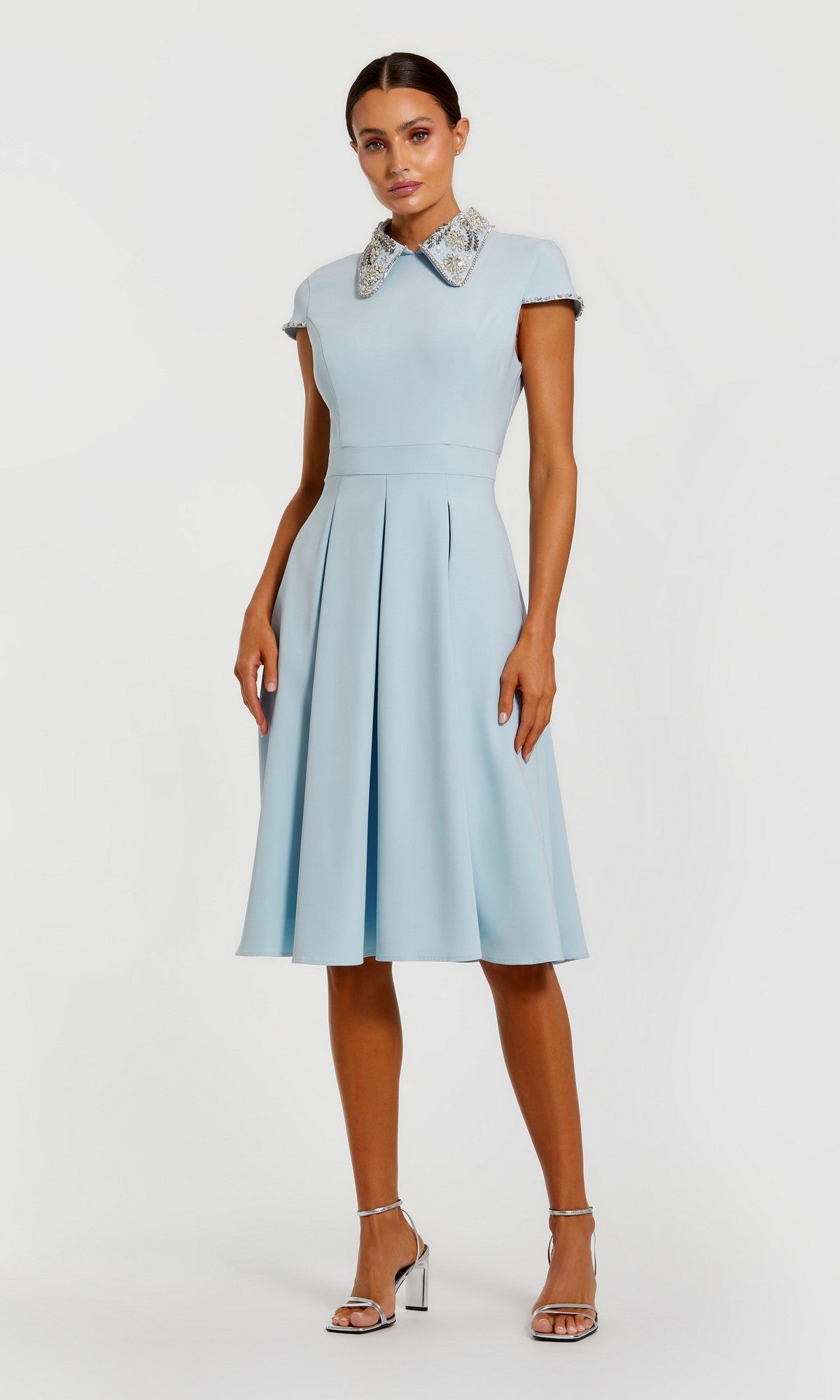 A woman in the powder blue Midi Wedding Guest Dress: Mac Duggal 2313 with cap sleeves, an A-line skirt, and a crystal-embellished collar pairs it with silver high-heeled sandals against a plain background.